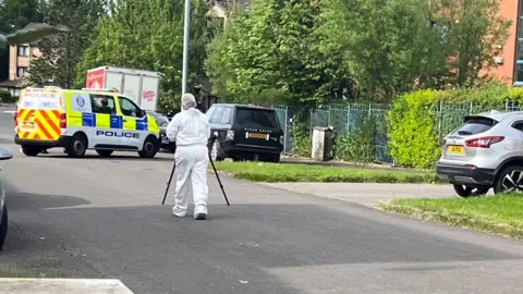 A forensics officer in white overalls on a street with a police van and parked cars 