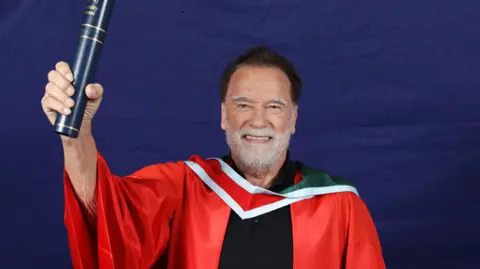 Arnold Schwarzenegger, a man with dark hair and a white beard, smiles while holding a navy and gold scroll holder in one hand. He is wearing a red university gown with a white and red collar