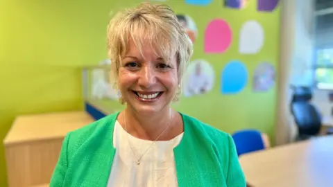 NIKKI FOX/BBC Sam Robinson looks at the camera wearing a bright green jacket. She has a white top on underneath it. She has short, blonde hair and is wearing earrings. She is smiling and is standing in the Big C office which is bright with a lime green wall and different colour teardrops showing photographs of people on the wall.