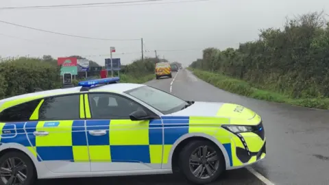 A police car with blue flashing lights is parked across the turning into Meaver Road, a two-lane single carriageway, lined with hedges and fields on a grey cloudy day