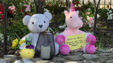 Stuart Woodward/BBC A close up shot of two soft toys propped up against railings with a flower bed behind. There is a white teddy with a heart-shaped nose and a pink unicorn. Next to the teddy is a yellow porcelain pot shaped like a chick with spring flowers in. A sign by the unicorn reads: "Rest in Peace, sorry for your loss, sending love and prayers".