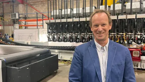 Alun stands at the end of a conveyor belt and in front of one of the robots. He has short light brown hair and wears a blue blazer over a white-striped shirt. 