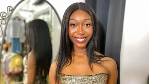 Lisa is standing in a charity shop wearing a gold ballgown with a black trim. Her long black hair is straight and worn down. You can see the back of her dress reflected in the mirror and she is standing in front of a changing room curtain smiling.