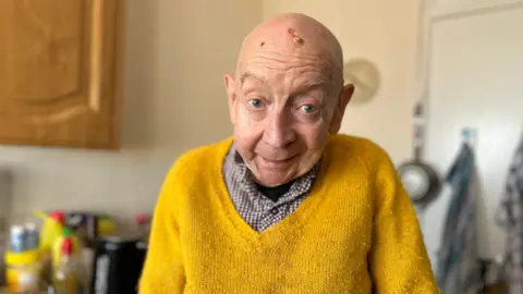 John Byrne who is bald standing in his kitchen with a shirt and yellow jumper