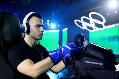 Getty Images A young man wearing white over-ear headphones sits in front of a videogame controller steering wheel in an arena setting. He wears an expression of concentration as two large screens glow green behind him and a large set of white Olympic rings lit in white hovers over the stage