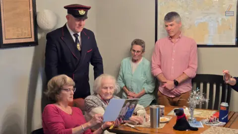 Luke Francis The family gathered around Emlyn Makofsky showing her the card from His Majesty the King. The card has a picture of King Charles and Queen Camilla on the front and Luke is stood behind Emlyn wearing his postmaster uniform. 