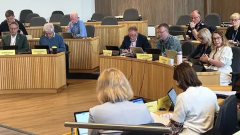 Carmelo Garcia A group of council members sit at desks at a Gloucestershire County Council meeting.