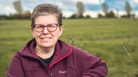 Lorna Vyse Lorna Vyse smiling at the camera. There is a field in the background. She is wearing glasses and a red fleece jacket with "Berghaus" branding on it