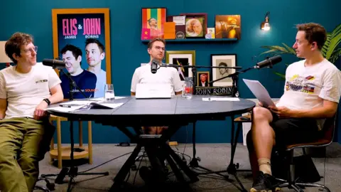 Elis & John in the podcast studio, sat around a table in front of microphones.
