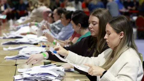 A woman in a cream jumper, with brown hair, looks at a white ballot paper , there is a woman with dark hair and a dark jumper beside her.