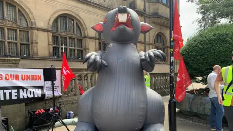 A giant inflatable grey rat is tethered in front of Sheffield Town Hall. In the background are trade union banners and flags.