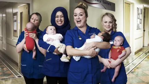 Four female midwives are standing together and smiling as they each hold a baby. They are in a hospital corridor, with a sign behind them saying 'delivery suites'.