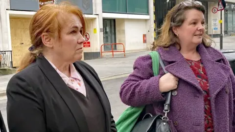 PA Media Two women stand on a street, looking to the right at a person off-camera. The woman on the left, Joanne Billington, has ginger hair tied back, and is wearing a pink shirt, black jumper, and black jacket. The woman on the right wears a red top and a purple coat.