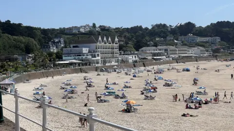 A view of a light. sandy beach on Jersey. There's people sat on the beach on blankets and umbrellas. There are buildings lining the seafront as well as trees surrounding them. 