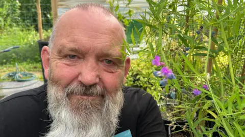 Volunteer John Barnard is crouching next to a marsh pea flower which is a purple and pink coloured bloom. He is bearded and smiling.