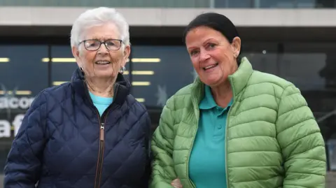 James Chapelard Two women standing outdoors side by side, smiling at the camera. One has white hair, glasses and a navy blue coat. The other has black hair, a green jacket and a blue polo shirt.