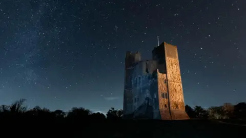 Orford Castle on a clear night. Stars can be seen in the sky above. The castle is lit by an orange glow.