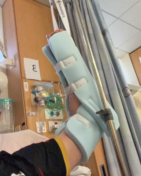 Adam Lang A photo of Adam's arm wrapped in a light blue protective sponge layer from the elbow up. His arm is propped upright and looks like it is attached to a pulley. A wall with hospital equipment attached can be seen in the background. 