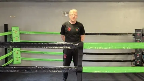 A man standing a boxing ring. He is wearing a black T-shirt and trousers and has his hands behind his back. The boxing ring is green and black.