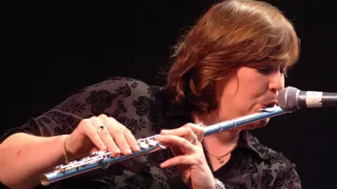 Getty Images A woman is onstage playing a flute. She has short brown hair and is wearing a watch, a bracelet and rings on both hands. 