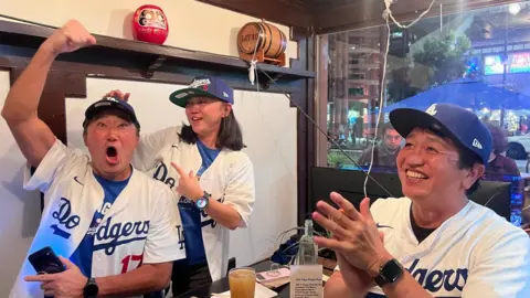 Three people cheer at a table inside a bar wearing Dodgers jerseys. 