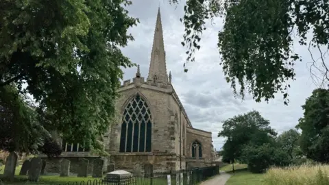 Harry Stevens/BBC A large stone church, with trees and bushes around it. There is a graveyard in front of the building. It has a large spire.