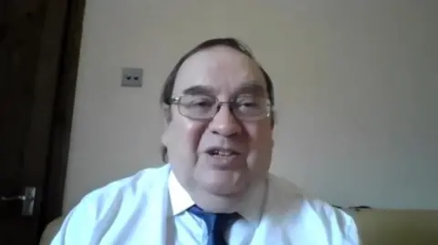 Chris Boden speaking on a video call. He has short black hair swept to one side and glasses, and is wearing a white shirt and blue tie. Behind him is a white wall and door.