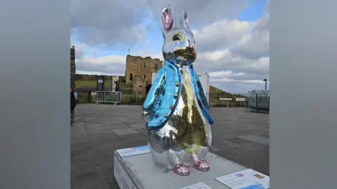 St Oswald's Hospice A Peter Rabbit sculpture covered in blue, silver and yellow sequins standing in front of a ruined priory. It is wearing Peter Rabbit's signature blue jacket with pink shoes. Tynemouth Priory is behind it with a black fence in front. The sky is blue with grey and white clouds.