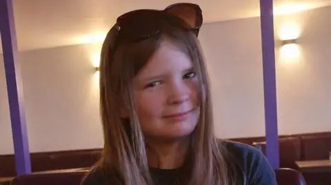 Faith Curran has shoulder-length blonde hair and has sunglasses on top of her head. She is smiling at the camera 