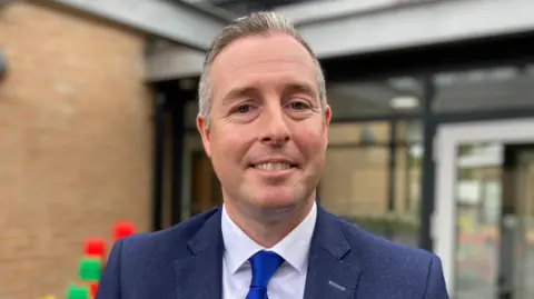 A close-up image of Paul Givan. He has short, greying hair and is standing outside a light coloured brick builing. He is smiling at the camera. He is wearing a navy suit with a blue tie and a white collared shirt.