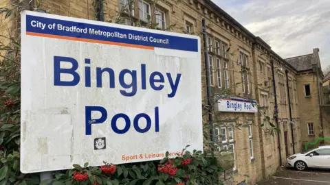 BBC Close-up shot of a white street sign with Bingley Pool in large, blue lettering and a yellow Yorkshire stone building in the background.   