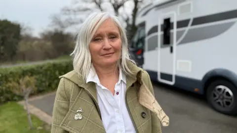 Steve Hubbard/BBC Joanne Fenton dengan rambut pirang panjang dan jaket berwarna batu dengan kemeja putih di bawahnya. Dia melihat kamera. Dia berada di samping motor putih dengan detail abu -abu dan hitam.