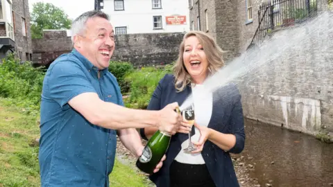 National Lottery A couple celebrate by shaking up a bottle of champagne next to a river.
