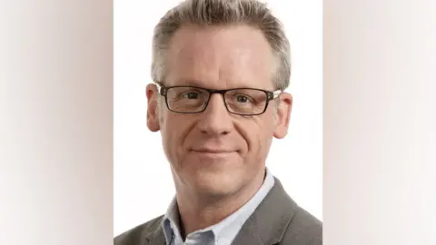 Mark Miller A man is looking at a camera with a white background behind him. He is in a shirt and blazer and is wearing glasses.