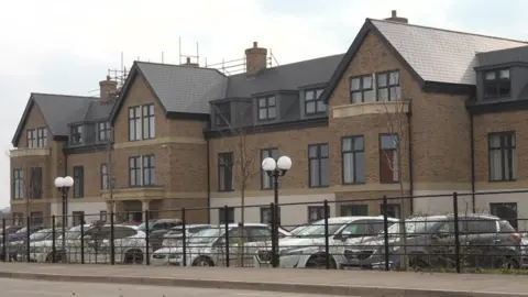 A modern brick care home with large windows and dark slate-style roofing. Several cars are parked in front of the building and are visible behind a black metal fence.