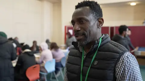 A man with a black body warmer, cheque shirt and green lanyard with short cropped hair and a dark but greying beard stands in front of a packed room of people sitting at a desk behind him.