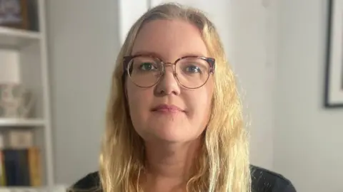 Emily Barley is sitting on a sofa in her lounge and she is holding a piece of paper which has two small handprints printed on it. Ms Barley has long blonde hair and is wearing glasses and a black top. There are framed photographs on the shelves to her right.