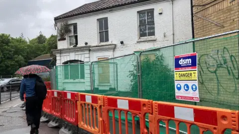 Owen Sennit/BBC A woman with an umbrella walks past in front of a derelict house on the corner of the street, with security railings around it.