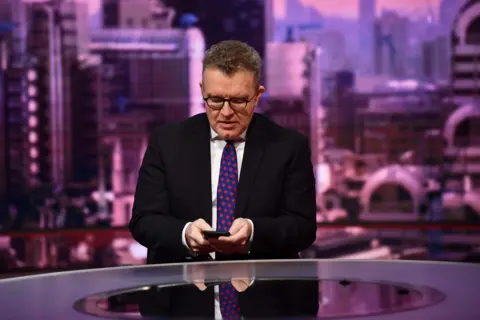 Tom Watson looks down at his phone in the BBC studios. He wears a dark suit jacket, white shirt and patterned tie. She wears glasses and has short hair 