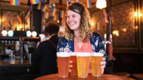 Getty Images A young woman smiles while holding pints of beer in a pub