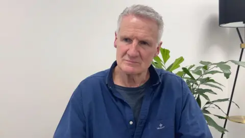 BBC Deputy Tom Binet is wearing a blue shirt and a black t-shirt. There is a plant behind him and a lamp 