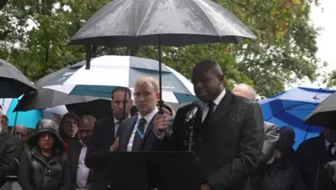 David Lammy is speaking at a podium, holding an umbrella to shield himself from the rain. Other people holding umbrellas stand behind him.