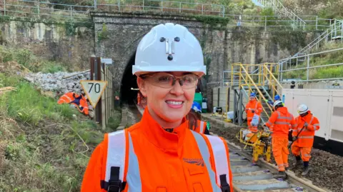 Charlotte Tyler, Network Rail project manager, looks towards the camera wearing a bright orange high-vis, behind her is a rail track and various workers in the same attire, with the Honiton tunnel seen in the background. 