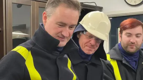 The photo shows three people wearing protective gear - which are black jackets with yellow stripes down either arm. The man in the middle is wearing a safety helmet. They are looking down at something in front of them. They're standing inside a room with cabinets and a wall clock in the background.