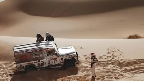 A.S.O./Aurélien Vialatte A land rover defender stuck in deep sand inside a sand dune. The driver of the car is stood with hands on hips outside of the car whilst two other people help to get the car out of the sand.