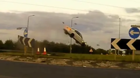 A white car caught mid‑air above a roundabout, tilted upward with debris scattered below, surrounded by road signs and traffic cones under a cloudy sky.