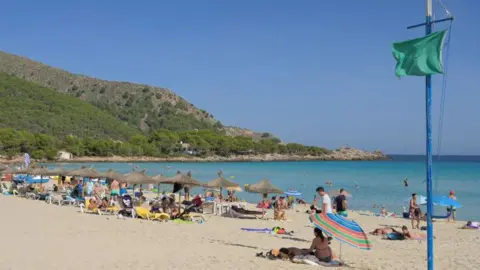 A sandy beach with sunbathers, swimmers, a green flag on a pole and straw beach umbrellas. A rocky spur is in the background at the far end of a bay.