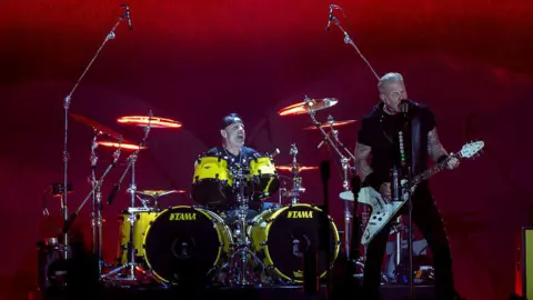 The rock band Metallica on stage - singer and guitarist James Hetfield sings into a microphone while drummer Lars Ulrich plays behind him.