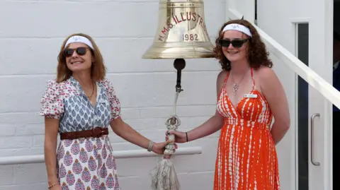 PA Media Amanda Thorpe, wife of former England cricketer Graham Thorpe and their daughter Emma Thorpe ring the bell ahead of the day's play on 'A Day for Thorpey' in support of Mind charity Action. Amanda wears a blue, red and white paisley pattern dress, and Emma wears an orange and white sundress. Both are wearing white headbands and both are holding the clapper of a bell from the ship HMS Illustrious.