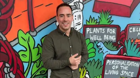 Curtis Silver A man with short dark hair in a green shirt stands in front of a mural with the words "plants for well being" on it